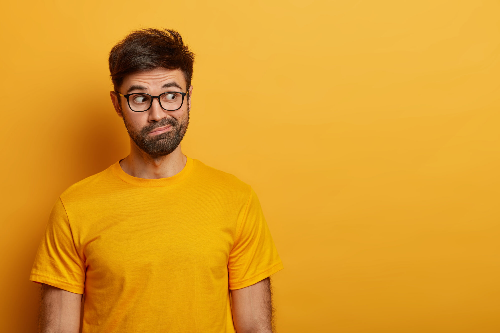 Photo of hesitant bearded man looks aside, smirks face and has puzzled expression, tries to decide something, dressed in casual yellow t shirt, poses over vibrant studio wall, wonders what he sees